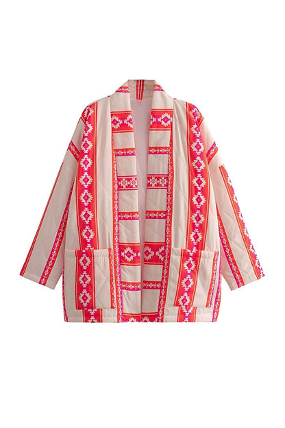 A woman wearing a relaxed, open-front kimono jacket featuring a vibrant pink and orange geometric Aztec print over a cream base, styled over a crochet crop top and shorts.