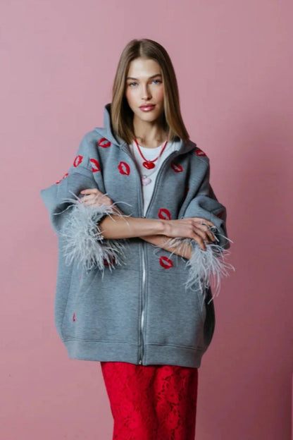 Grey - A woman wearing an oversized grey zip-up hoodie featuring a red lip print and dramatic grey feather cuffs, styled over a white top and red textured skirt.