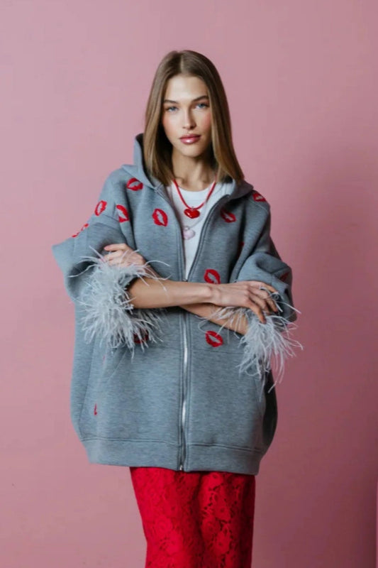 Grey - A woman wearing an oversized grey zip-up hoodie featuring a red lip print and dramatic grey feather cuffs, styled over a white top and red textured skirt.