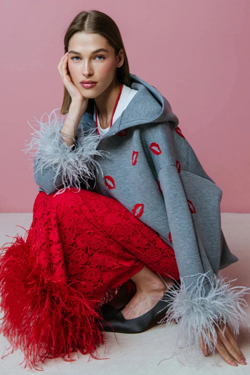 Grey - A woman wearing an oversized grey zip-up hoodie featuring a red lip print and dramatic grey feather cuffs, styled over a white top and red textured skirt.