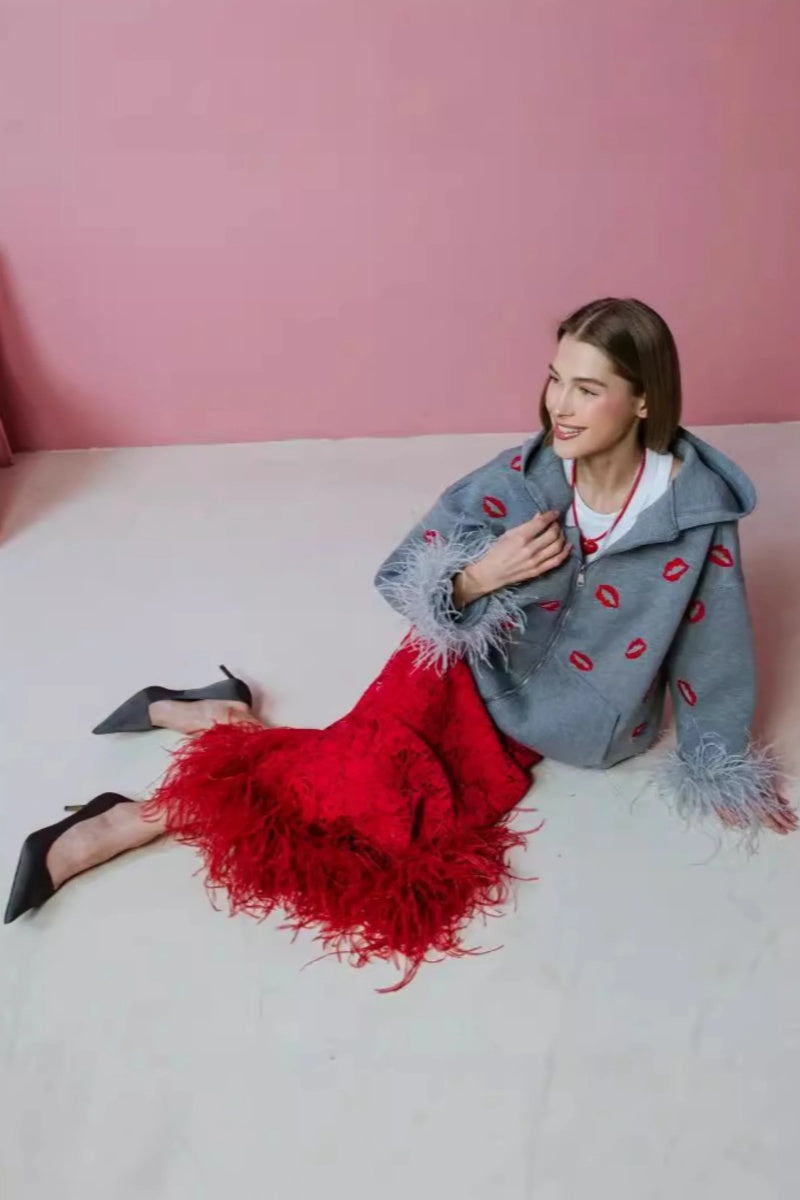 Grey - A woman wearing an oversized grey zip-up hoodie featuring a red lip print and dramatic grey feather cuffs, styled over a white top and red textured skirt.
