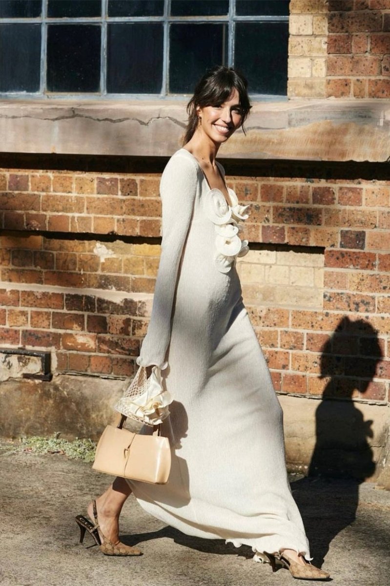 White - A woman wearing a long-sleeved, white ribbed knit maxi dress featuring a deep plunging neckline decorated with large 3D floral appliqués.
