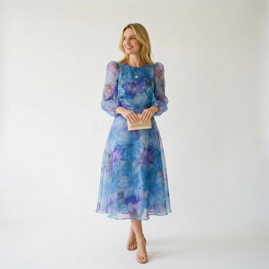 Blue - A woman stands wearing a blue and purple floral print chiffon midi dress with long sheer puff sleeves, a high neckline, and an A-line skirt, holding a gold clutch.