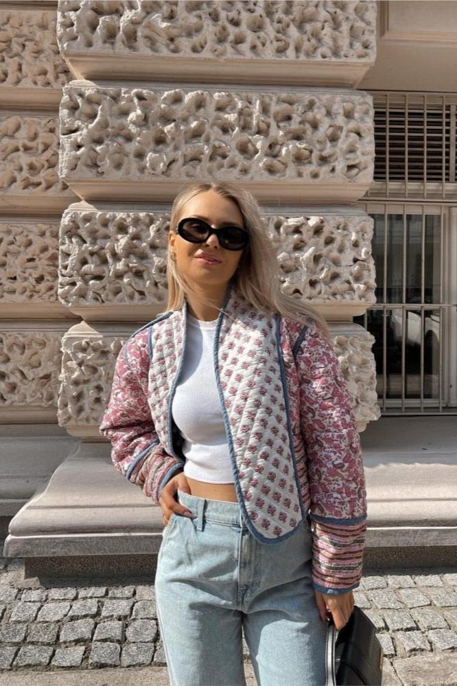 Women's open-front quilted jacket featuring a pink and white floral block print, contrasting blue edge binding, and diamond stitching, worn over a white tee and light wash jeans.