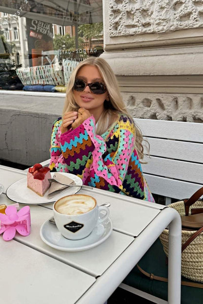 A woman taking a mirror selfie wearing an oversized, multicolour patchwork crochet cardigan over a white top and blue jeans, holding a woven straw tote bag.