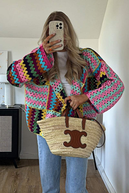 A woman taking a mirror selfie wearing an oversized, multicolour patchwork crochet cardigan over a white top and blue jeans, holding a woven straw tote bag.