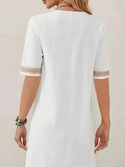 White - A woman wearing a white, knee-length shift dress made of textured linen-like fabric, featuring intricate beige crochet lace trim along the deep V-neckline and elbow-length sleeve cuffs.