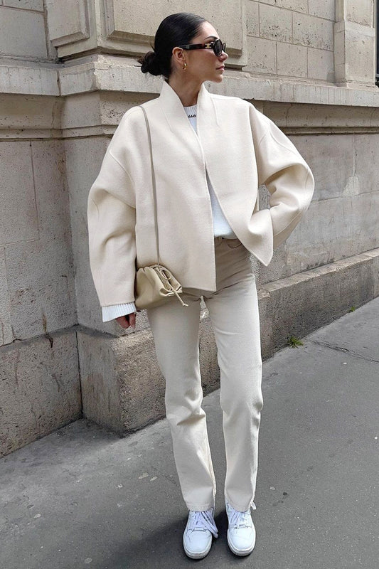 Woman wearing an oversized, collarless, open-front cropped jacket in a light cream colour, styled over a white knit sweater and cream jeans.