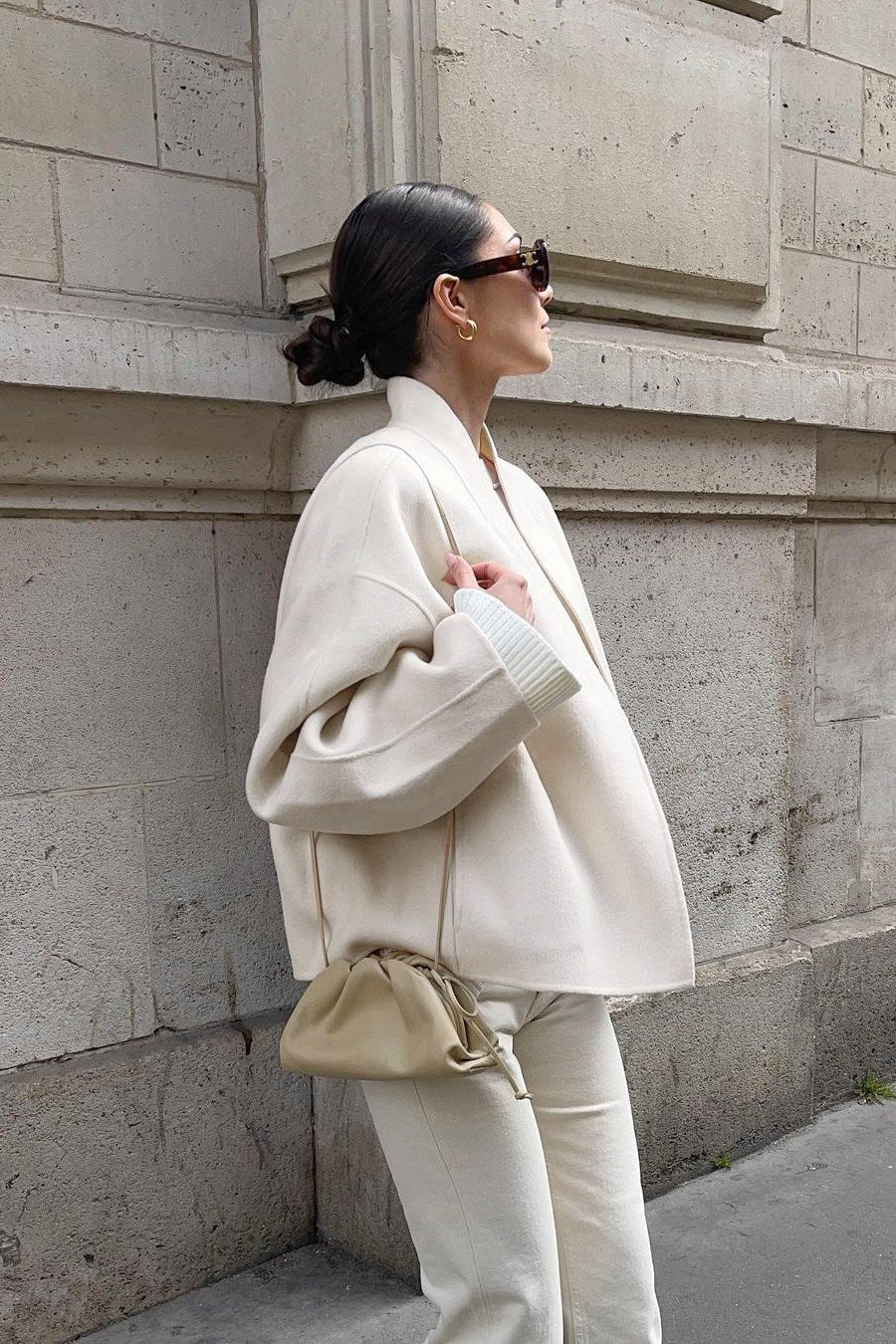 Woman wearing an oversized, collarless, open-front cropped jacket in a light cream colour, styled over a white knit sweater and cream jeans.