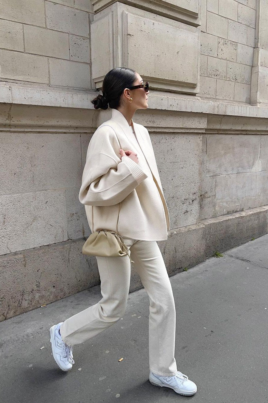 Woman wearing an oversized, collarless, open-front cropped jacket in a light cream colour, styled over a white knit sweater and cream jeans.
