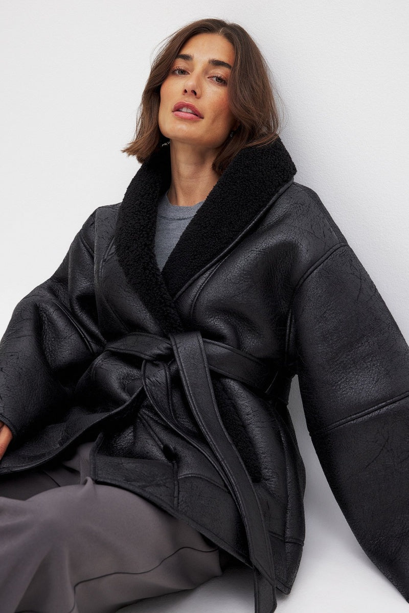 Black - A woman wearing a black, oversized faux leather coat with a plush faux shearling lapel, cinched at the waist with a self-tie belt, standing against a wood-paneled wall.