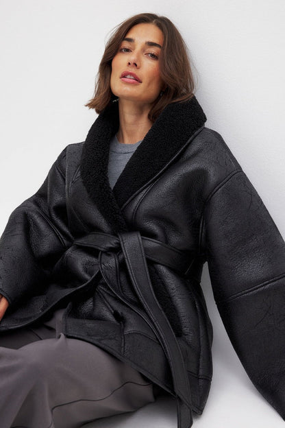 Black - A woman wearing a black, oversized faux leather coat with a plush faux shearling lapel, cinched at the waist with a self-tie belt, standing against a wood-paneled wall.