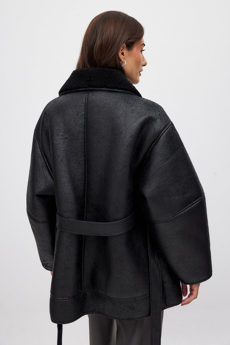 Black - A woman wearing a black, oversized faux leather coat with a plush faux shearling lapel, cinched at the waist with a self-tie belt, standing against a wood-paneled wall.