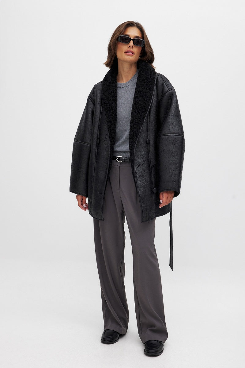 Black - A woman wearing a black, oversized faux leather coat with a plush faux shearling lapel, cinched at the waist with a self-tie belt, standing against a wood-paneled wall.