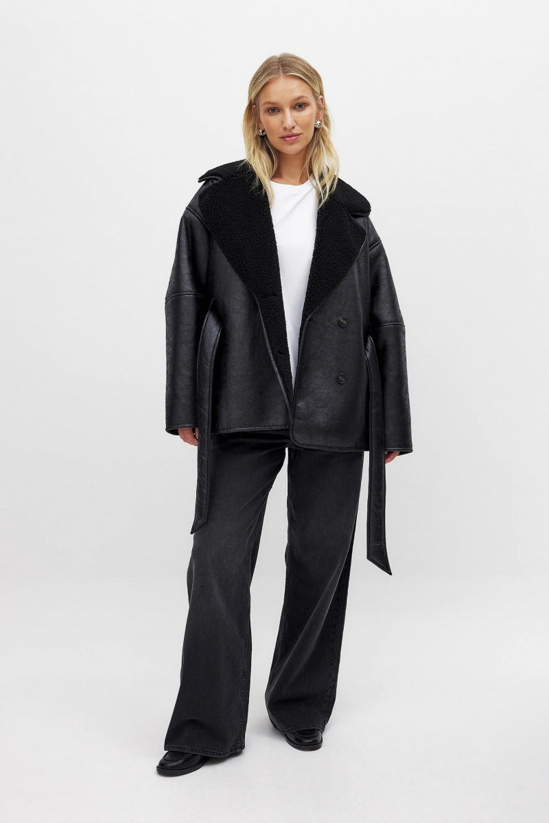 Black - A woman wearing a black, oversized faux leather coat with a plush faux shearling lapel, cinched at the waist with a self-tie belt, standing against a wood-paneled wall.