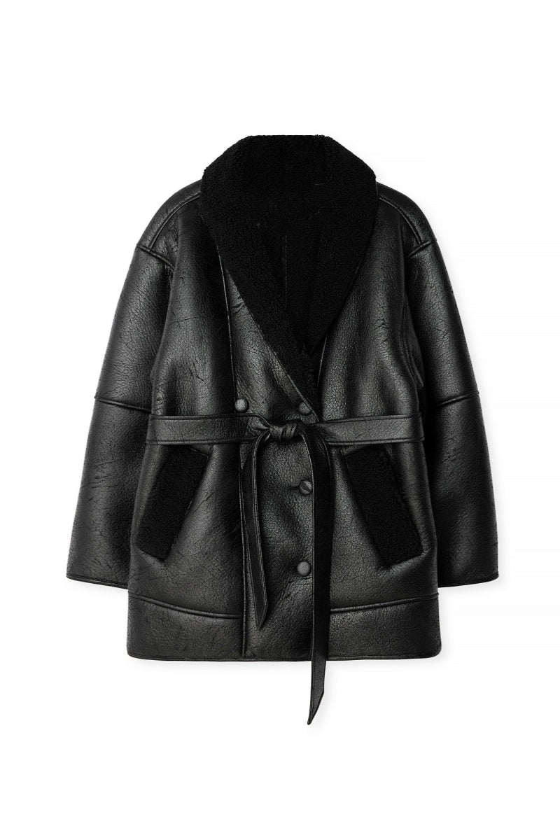 Black - A woman wearing a black, oversized faux leather coat with a plush faux shearling lapel, cinched at the waist with a self-tie belt, standing against a wood-paneled wall.