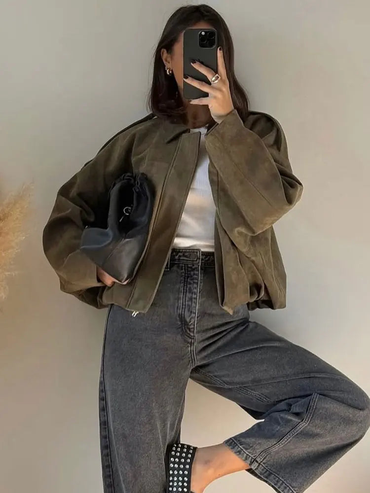 A woman taking a mirror selfie wearing an oversized, olive green faux-suede jacket with dropped shoulders, styled over a white top and dark grey jeans.