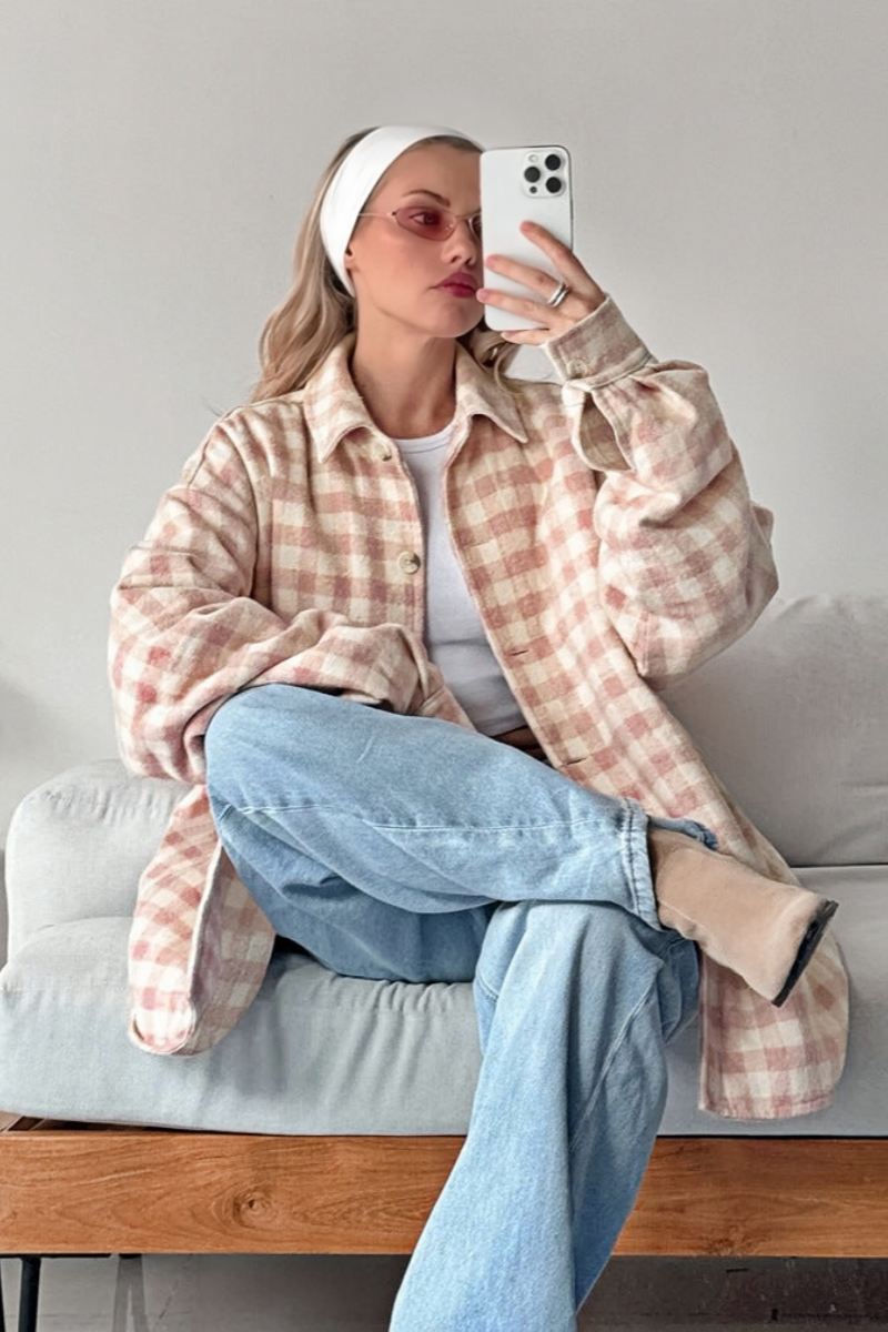 A woman taking a mirror selfie wearing an oversized, light pink and white plaid shacket over a white crop top and light wash denim jeans.
