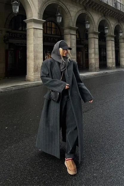 A woman walking on a city street wearing an oversized, structured charcoal pinstripe maxi coat with padded shoulders, styled with a grey hood and dark trousers.