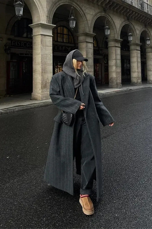 A woman walking on a city street wearing an oversized, structured charcoal pinstripe maxi coat with padded shoulders, styled with a grey hood and dark trousers.