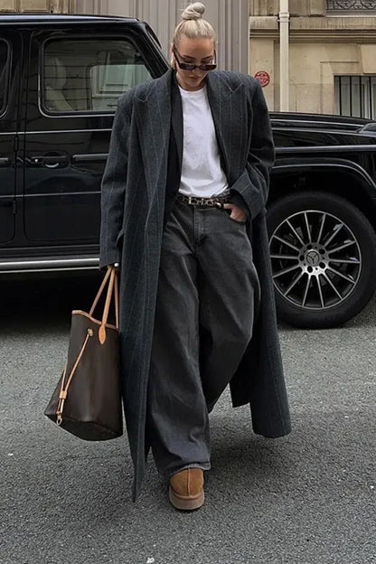 Grey - A woman walking on a city street wearing an oversized, structured charcoal pinstripe maxi coat with padded shoulders, styled with a grey hood and dark trousers.