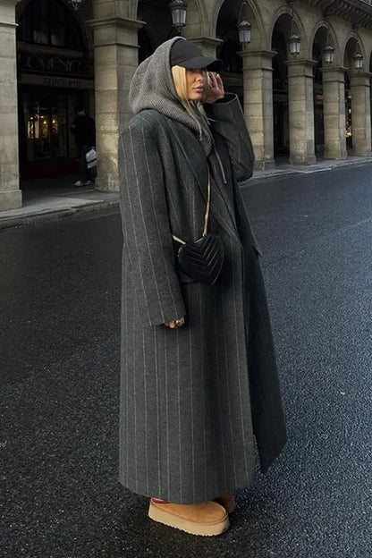 Grey - A woman walking on a city street wearing an oversized, structured charcoal pinstripe maxi coat with padded shoulders, styled with a grey hood and dark trousers.