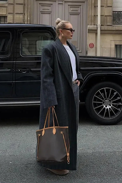 Grey - A woman walking on a city street wearing an oversized, structured charcoal pinstripe maxi coat with padded shoulders, styled with a grey hood and dark trousers.