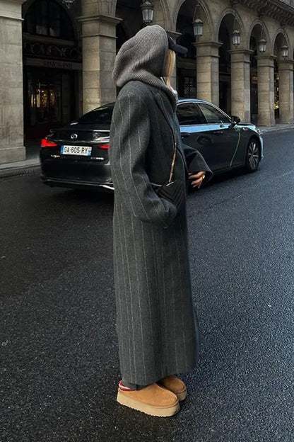 Grey - A woman walking on a city street wearing an oversized, structured charcoal pinstripe maxi coat with padded shoulders, styled with a grey hood and dark trousers.