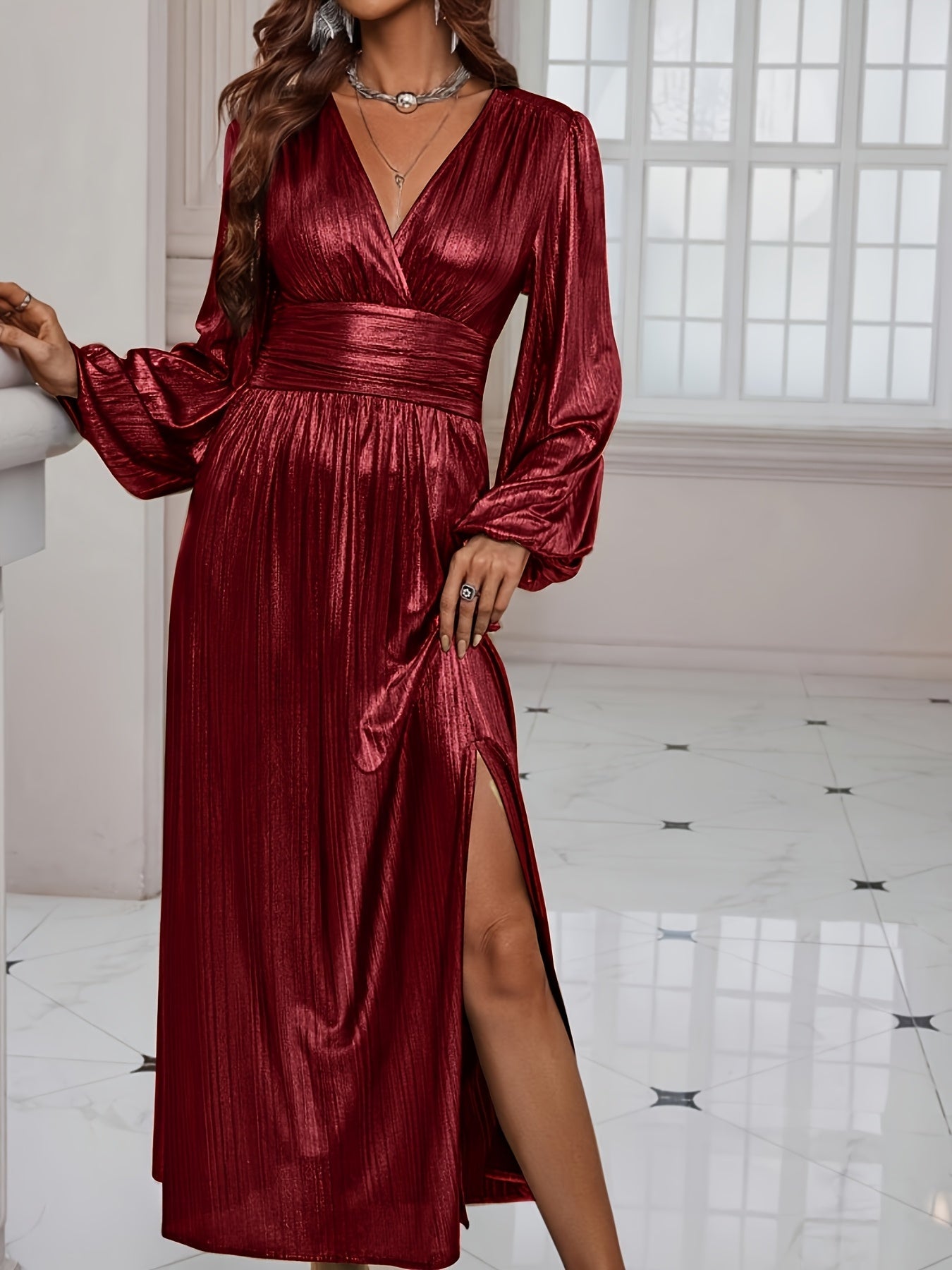 Woman wearing a shiny metallic pleated long sleeve maxi dress with a wrap bodice, banded waist, and high leg slit, standing in a formal indoor setting.