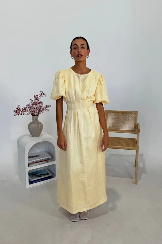 Butter Yellow - A woman wearing a pale yellow midi dress with short puff sleeves, a fitted waist, and a pleated skirt, standing in a minimalist studio setting.