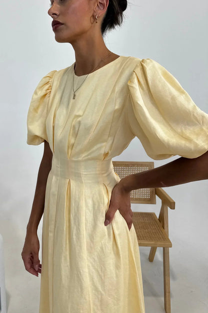 Butter Yellow - A woman wearing a pale yellow midi dress with short puff sleeves, a fitted waist, and a pleated skirt, standing in a minimalist studio setting.