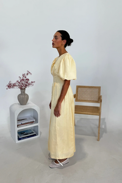Butter Yellow - A woman wearing a pale yellow midi dress with short puff sleeves, a fitted waist, and a pleated skirt, standing in a minimalist studio setting.