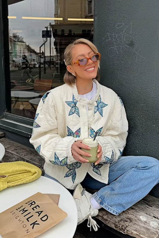 A woman sitting outdoors wearing a cream quilted jacket with blue star appliqués, styled with light wash jeans and sunglasses.