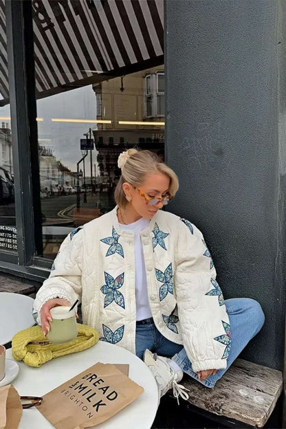 A woman sitting outdoors wearing a cream quilted jacket with blue star appliqués, styled with light wash jeans and sunglasses.