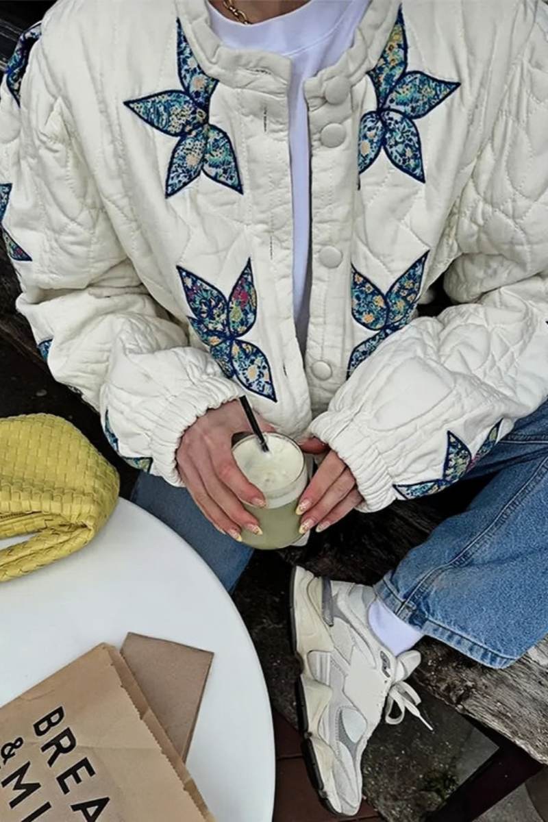 A woman sitting outdoors wearing a cream quilted jacket with blue star appliqués, styled with light wash jeans and sunglasses.