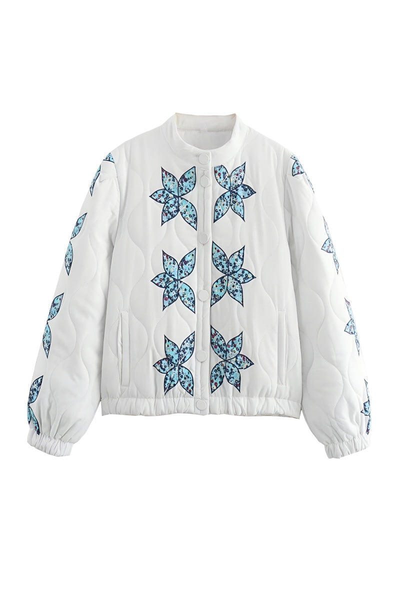 A woman sitting outdoors wearing a cream quilted jacket with blue star appliqués, styled with light wash jeans and sunglasses.