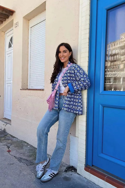 A woman stands outdoors wearing a relaxed-fit, hip-length quilted jacket featuring a bold blue and white geometric ikat print, styled open over a white crop top and straight-leg jeans.
