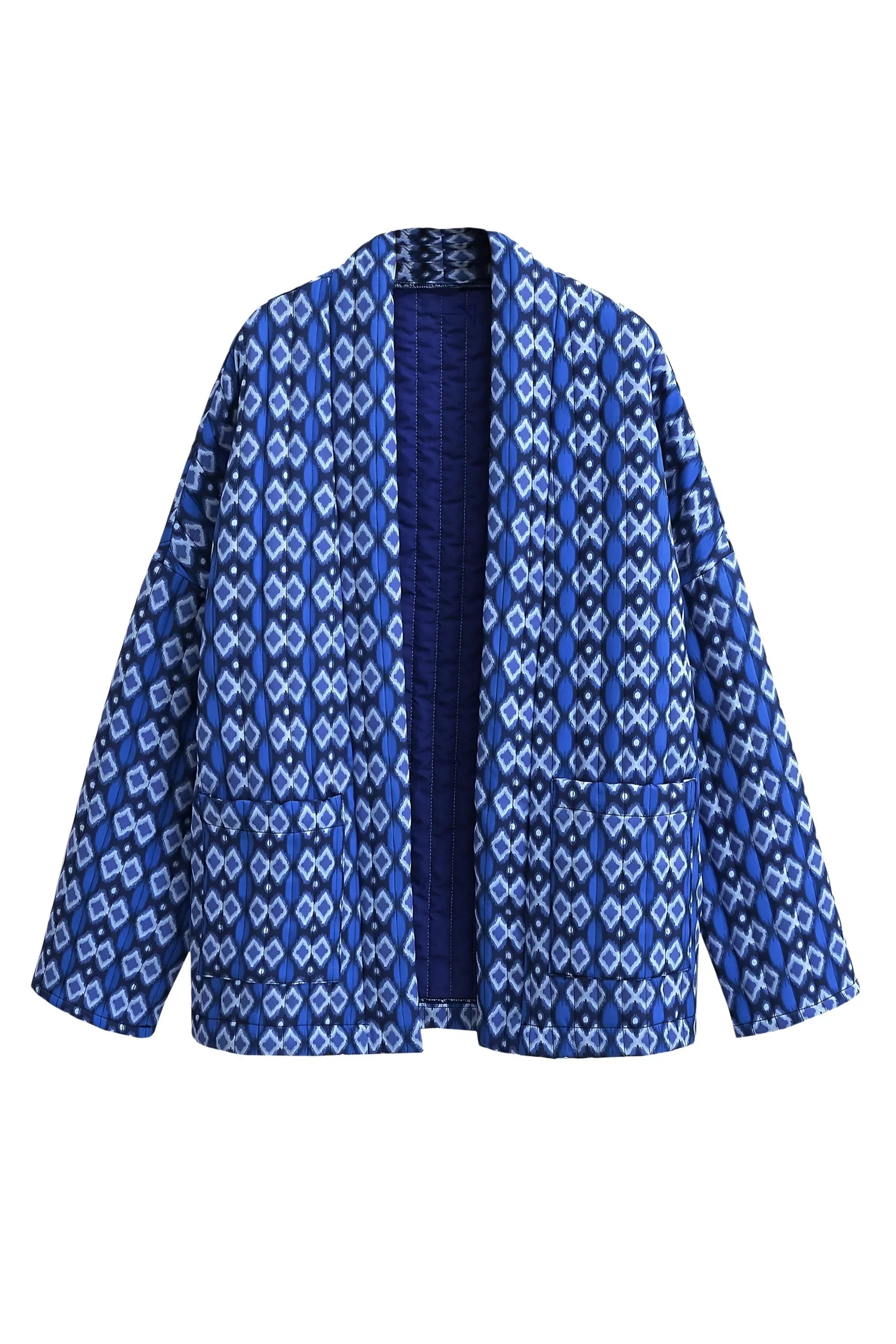 Blue - A woman stands outdoors wearing a relaxed-fit, hip-length quilted jacket featuring a bold blue and white geometric ikat print, styled open over a white crop top and straight-leg jeans.