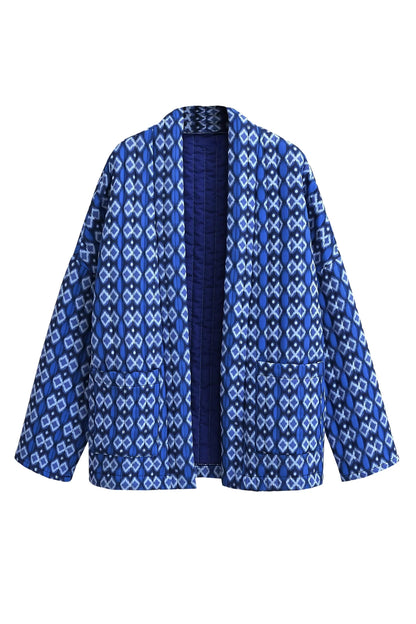 Blue - A woman stands outdoors wearing a relaxed-fit, hip-length quilted jacket featuring a bold blue and white geometric ikat print, styled open over a white crop top and straight-leg jeans.