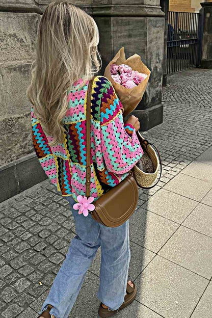 A woman taking a mirror selfie wearing an oversized, multicolour patchwork crochet cardigan over a white top and blue jeans, holding a woven straw tote bag.