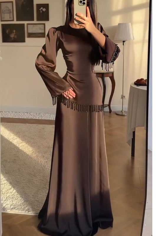 Brown - A woman wearing a floor-length chocolate brown satin maxi dress with long bell sleeves, featuring black beaded fringe detailing along the sleeve cuffs and an asymmetric hip seam.