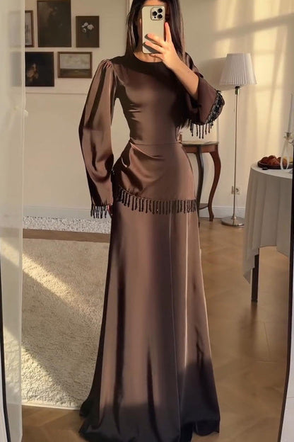 Brown - A woman wearing a floor-length chocolate brown satin maxi dress with long bell sleeves, featuring black beaded fringe detailing along the sleeve cuffs and an asymmetric hip seam.
