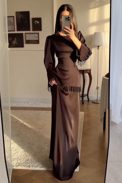 Brown - A woman wearing a floor-length chocolate brown satin maxi dress with long bell sleeves, featuring black beaded fringe detailing along the sleeve cuffs and an asymmetric hip seam.