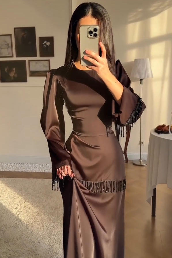 Brown - A woman wearing a floor-length chocolate brown satin maxi dress with long bell sleeves, featuring black beaded fringe detailing along the sleeve cuffs and an asymmetric hip seam.