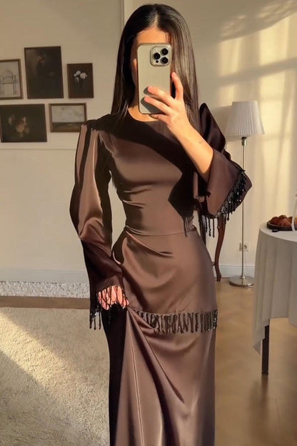 Brown - A woman wearing a floor-length chocolate brown satin maxi dress with long bell sleeves, featuring black beaded fringe detailing along the sleeve cuffs and an asymmetric hip seam.