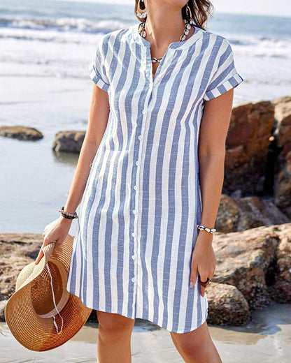 Blue / White - A woman wearing a blue and white vertically striped short-sleeve shirt dress with a split neckline, standing on a beach holding a straw sunhat.