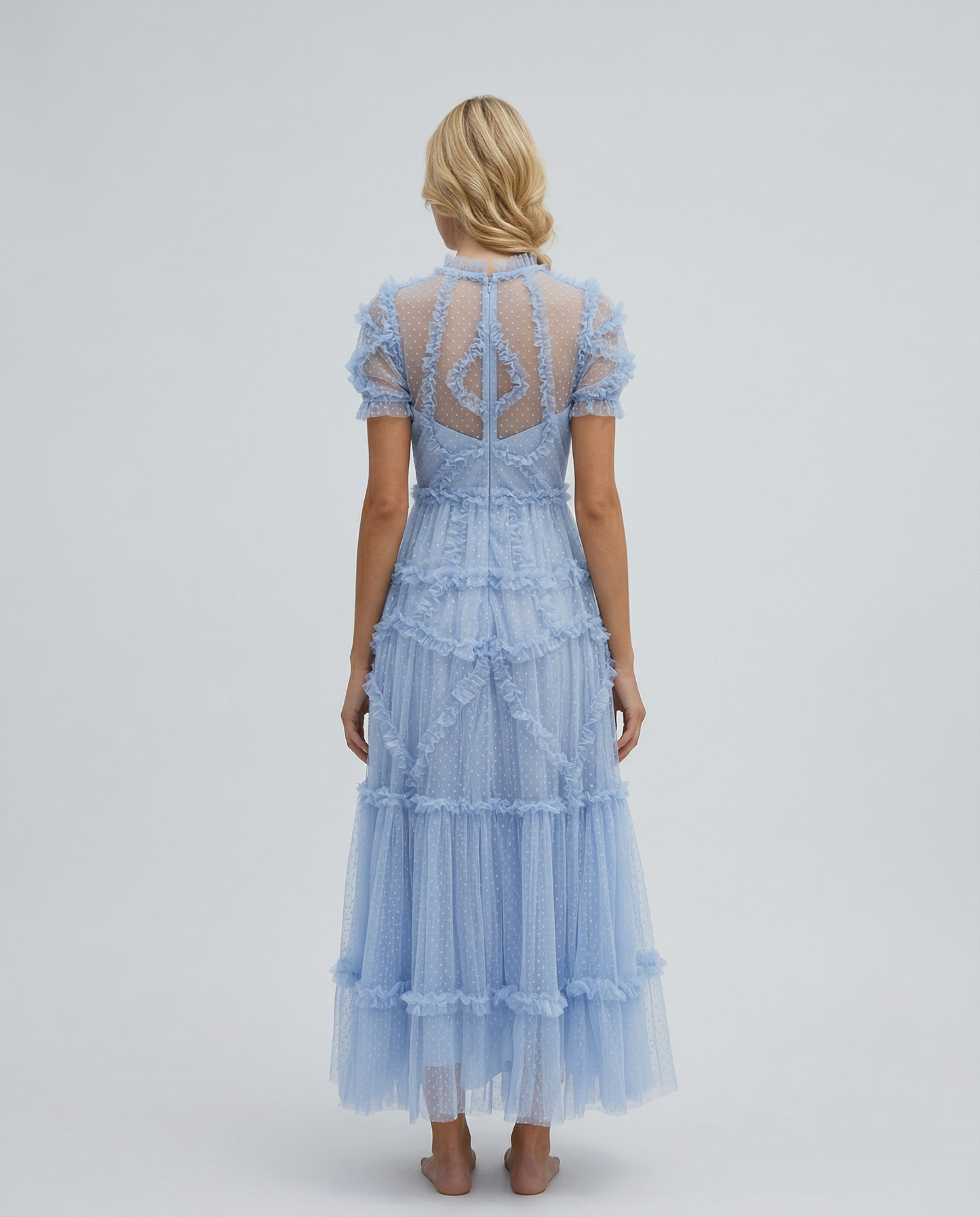 Blue - A woman wearing a light blue Swiss dot tulle maxi dress with a high neck, tiered skirt, and ruffled chevron detailing.