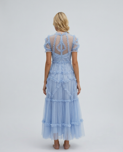 Blue - A woman wearing a light blue Swiss dot tulle maxi dress with a high neck, tiered skirt, and ruffled chevron detailing.
