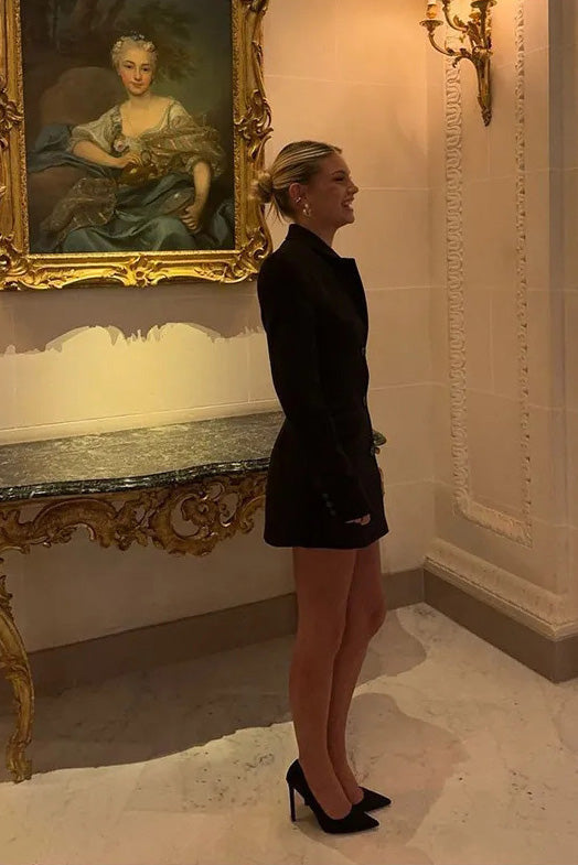Black - Model standing on stairs wearing a tailored long-sleeve mini blazer dress with front buttons and flap pockets.