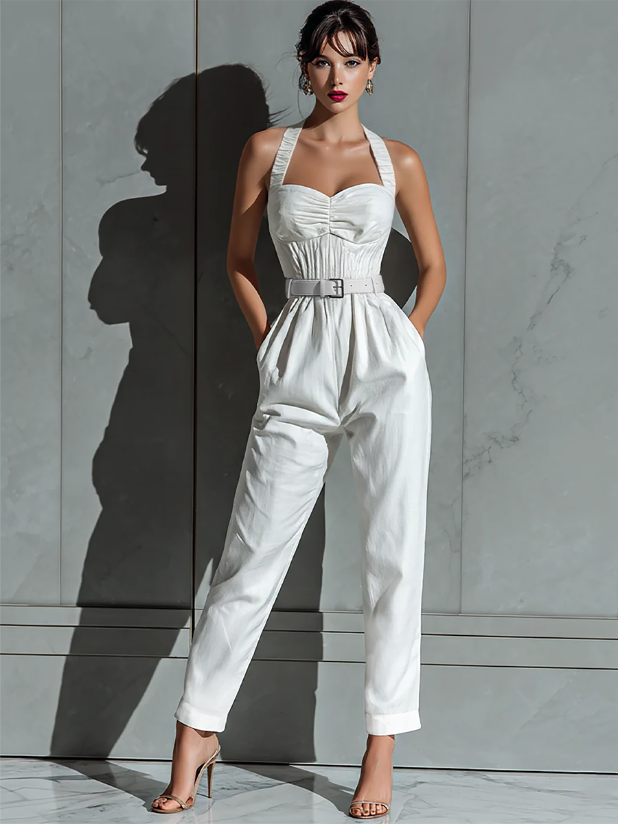 Ivory - A woman wearing a white textured linen halter-neck jumpsuit with a ruched bodice, a matching belted waist, and tapered trousers, standing against a grey marble wall.